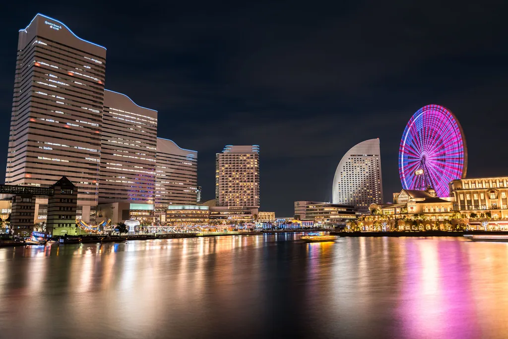 Tall buildings and a lit Ferris wheel reflected in a harbor at night